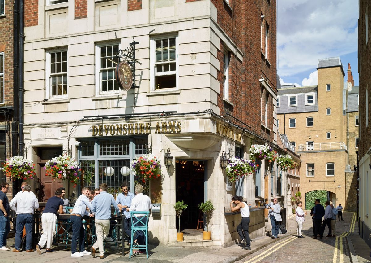 On a sunny day, after-work drinkers gather around high tables outside the Devonshire Arms, a white stone corner pub with gold lettering and colourful hanging ba