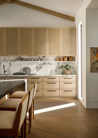 Kitchen cabinets that are made of oak with slatted upper cabinets and sleek, clean lower cabinets. The lower cabinets have long black handles, and opposite the cabinets is an island with bar stools.