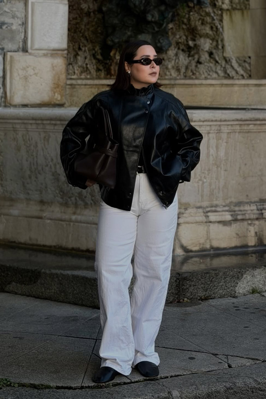 A woman standing outside in front of a building wearing black sunglasses with a black leather cape coat, white baggy wide-leg jeans, and black leather sock boots.