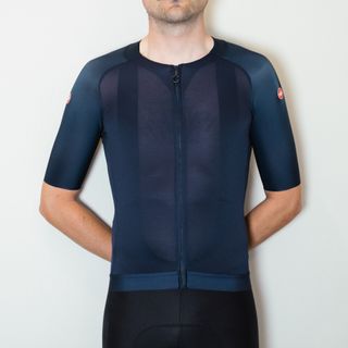 A white man in a navy blue cycling jersey and black cycling shorts stands against a white wall