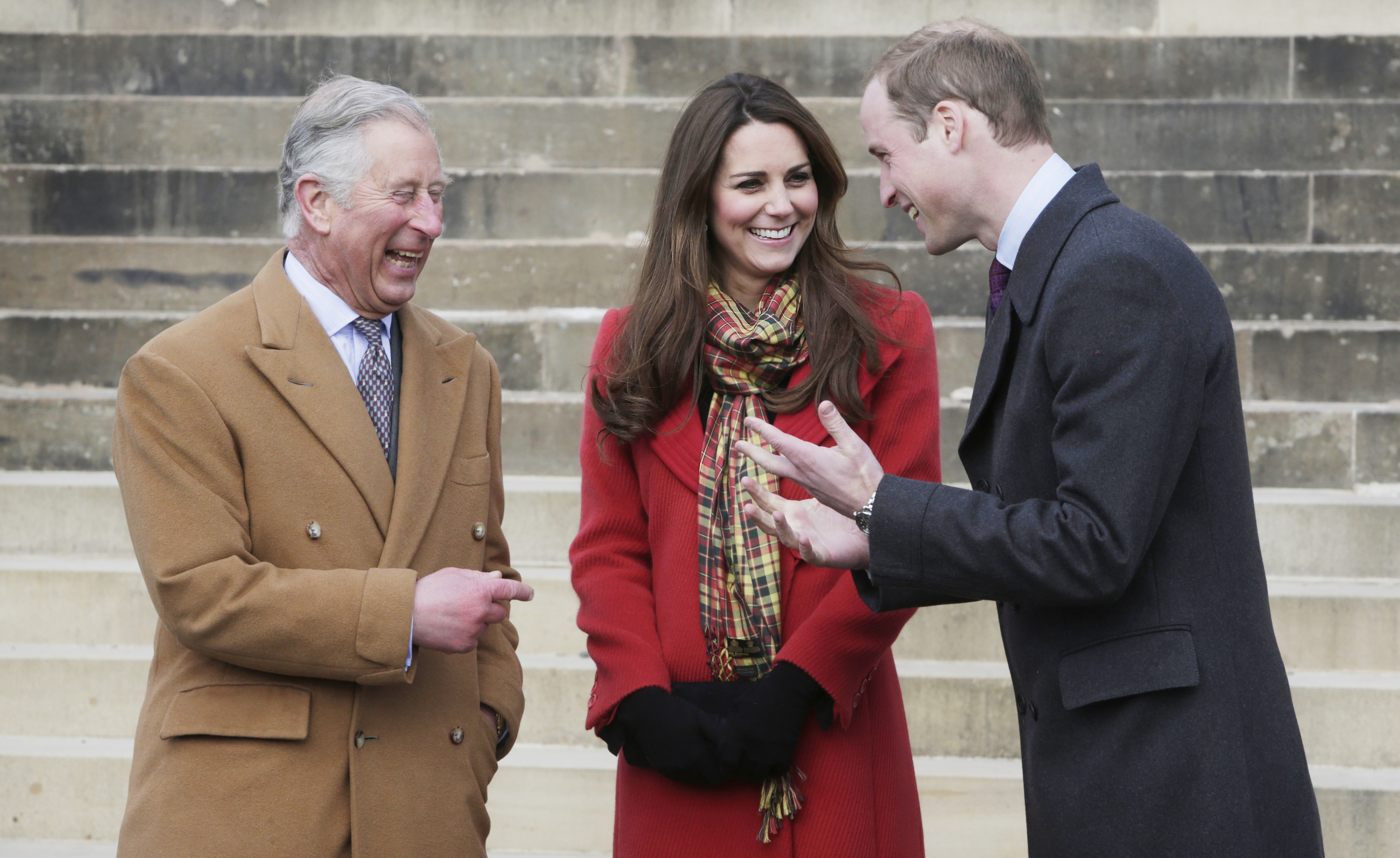 King Charles wears a brown overcoat and laughs alongside Kate Middleton, who wears a red coat with a tartan scarf, and Prince William