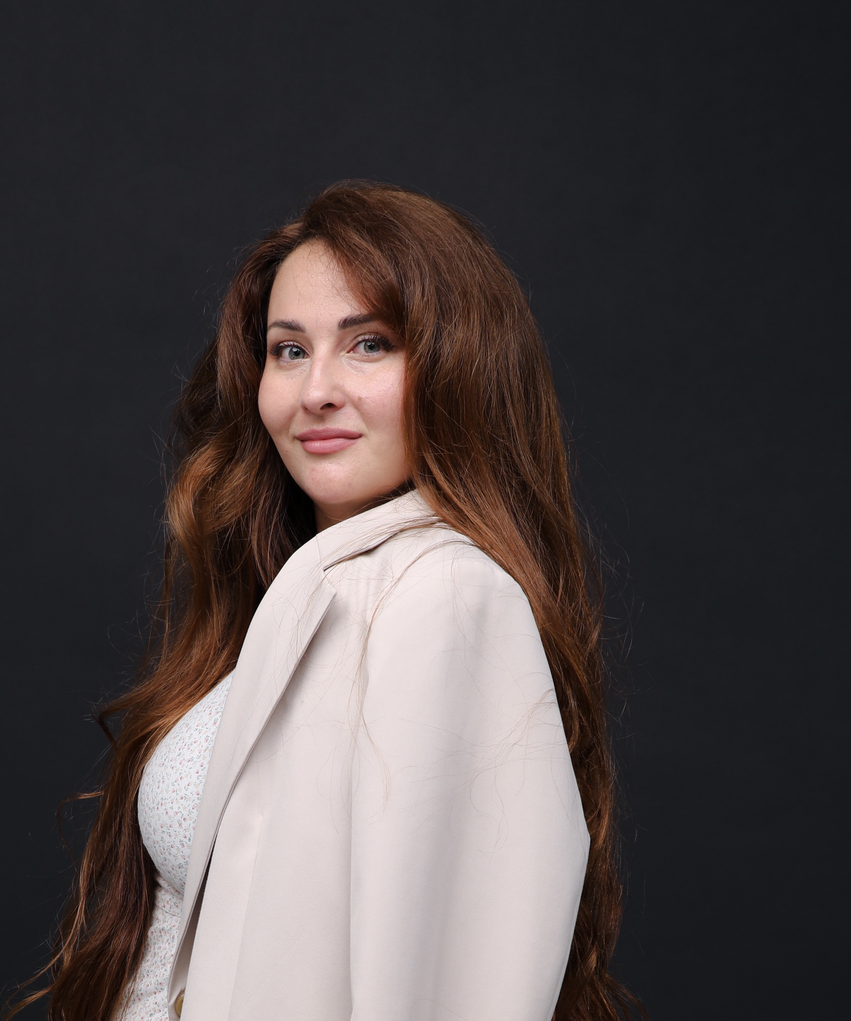 a headshot of a white woman with long, red hair wearing a cream blazer and posing in front of a dark background