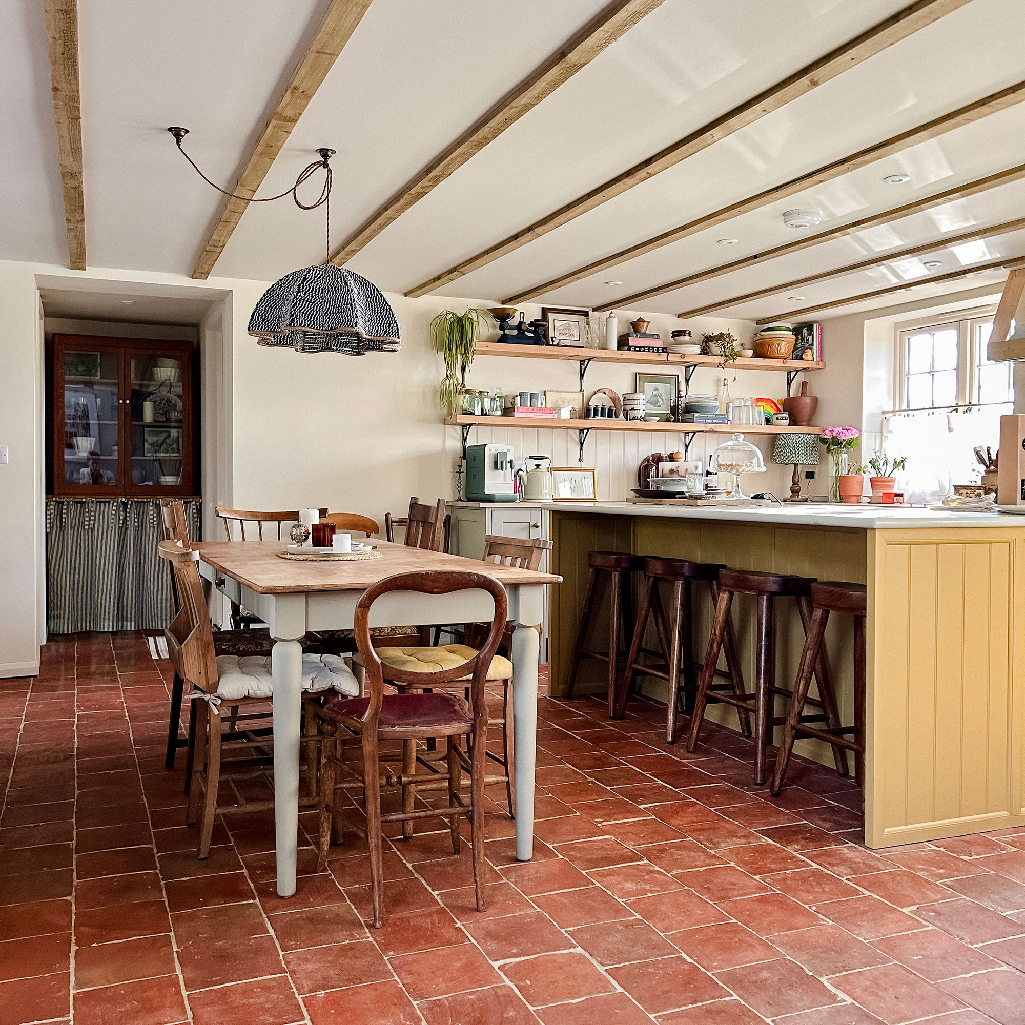 Kitchen dining area with yellow island, wooden dining table, pendant lighting, terracotta floors and wooden ceiling beams