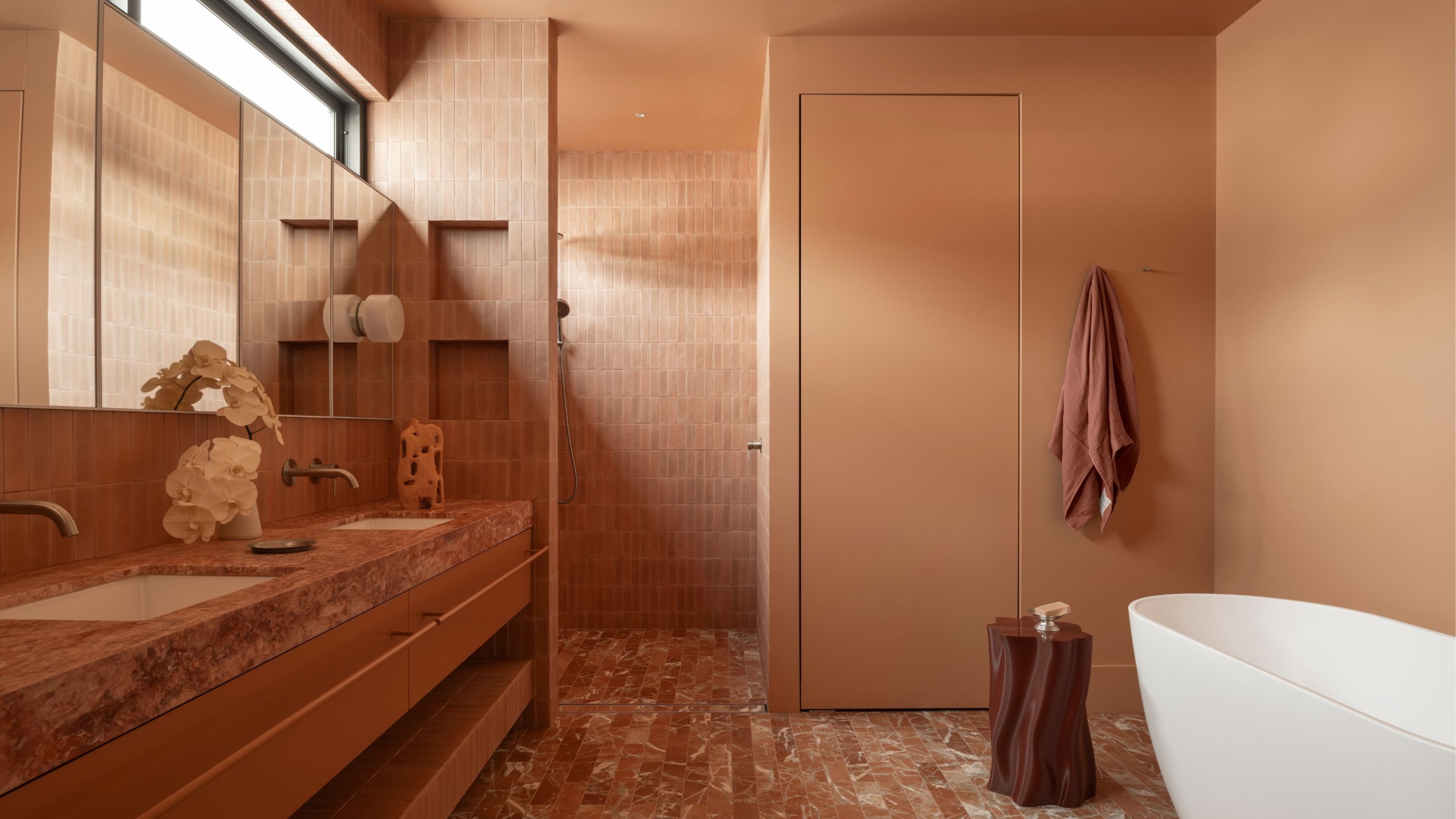 a terracotta color drenched bathroom with terracotta vanity and floor tiles, terracotta wall paint, white bath, terracotta towel hanging, stump side table, and mirrored cabinets