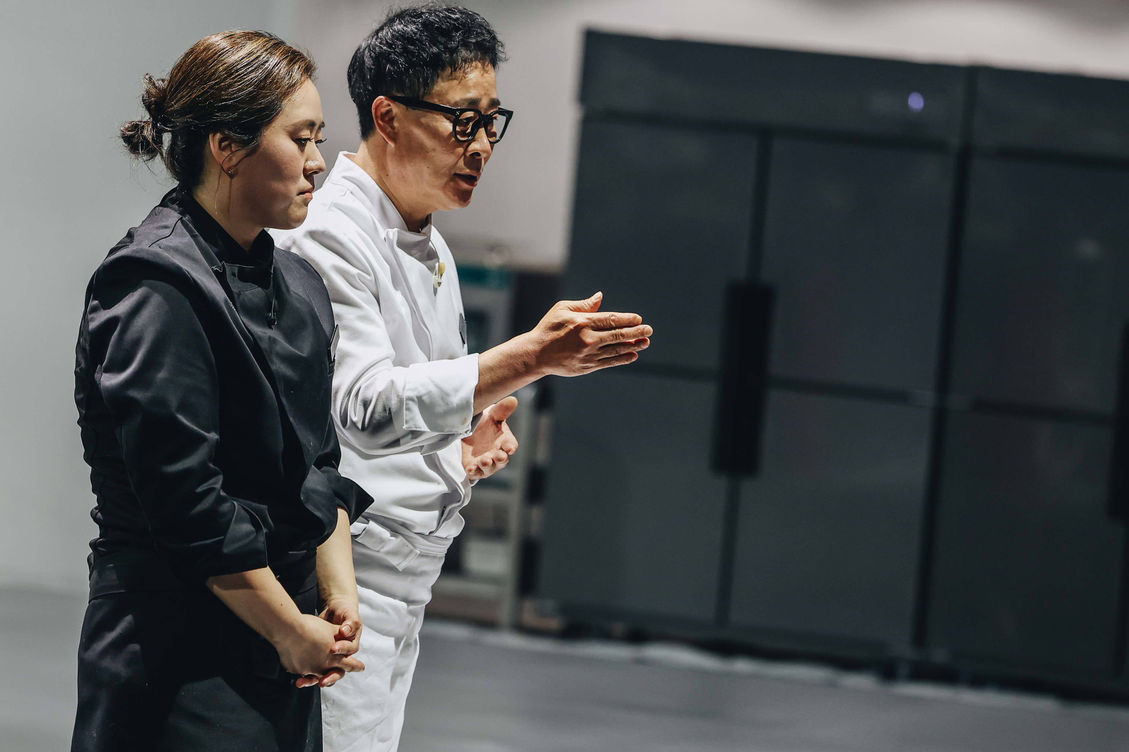 A female chef dressed in black (Brewmaster Yun) and a male chef dressed in white (Im Seong-keun.) They stand in an industrial kitchen while addressing someone offscreen, in 'Culinary Class Wars' season 2.