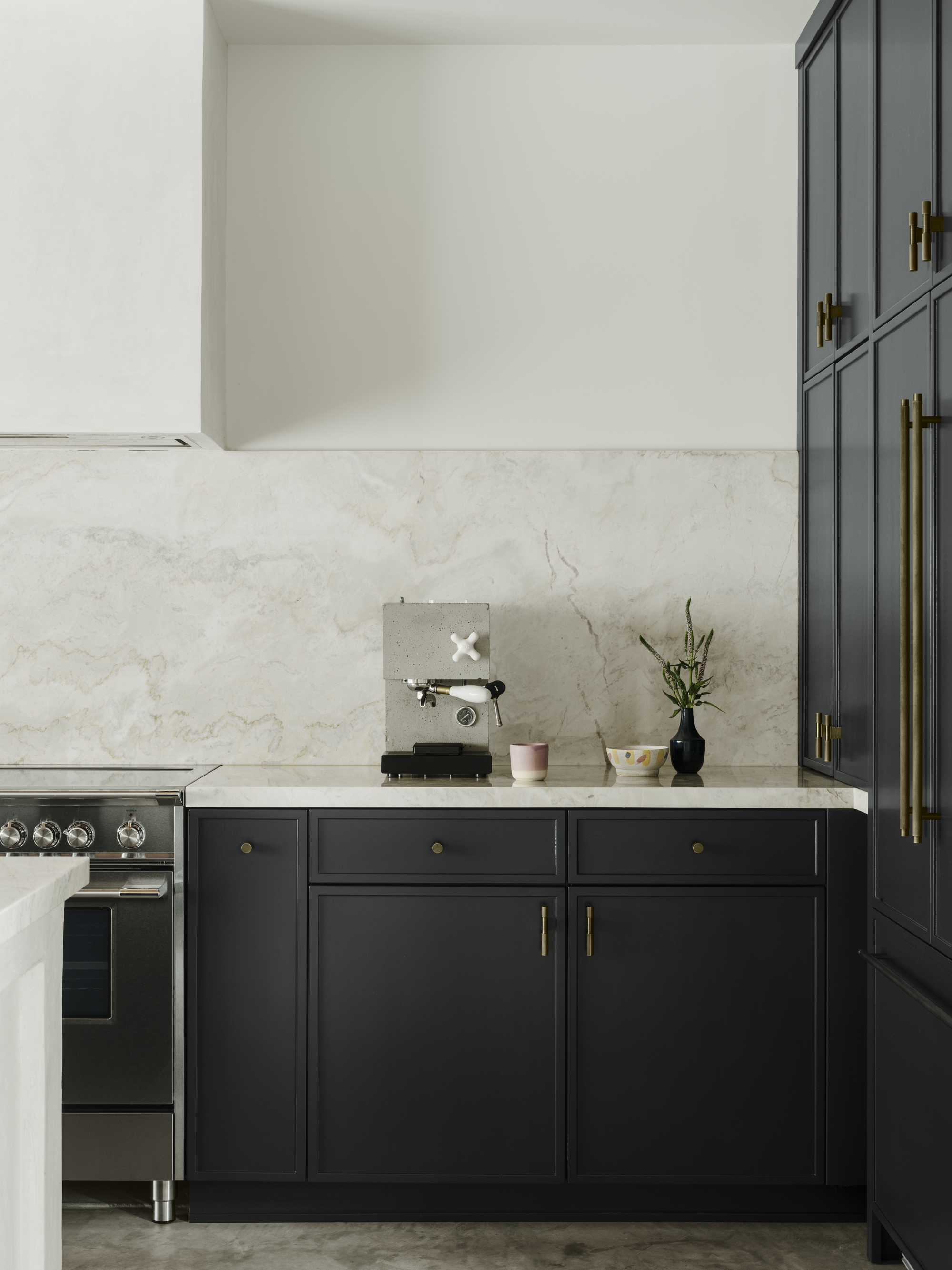 Image of a minimalist kitchen with white walls, navy cabinetry, and a white marble countertop and backsplash. There is a stylish, concrete espresso machine on the counter.