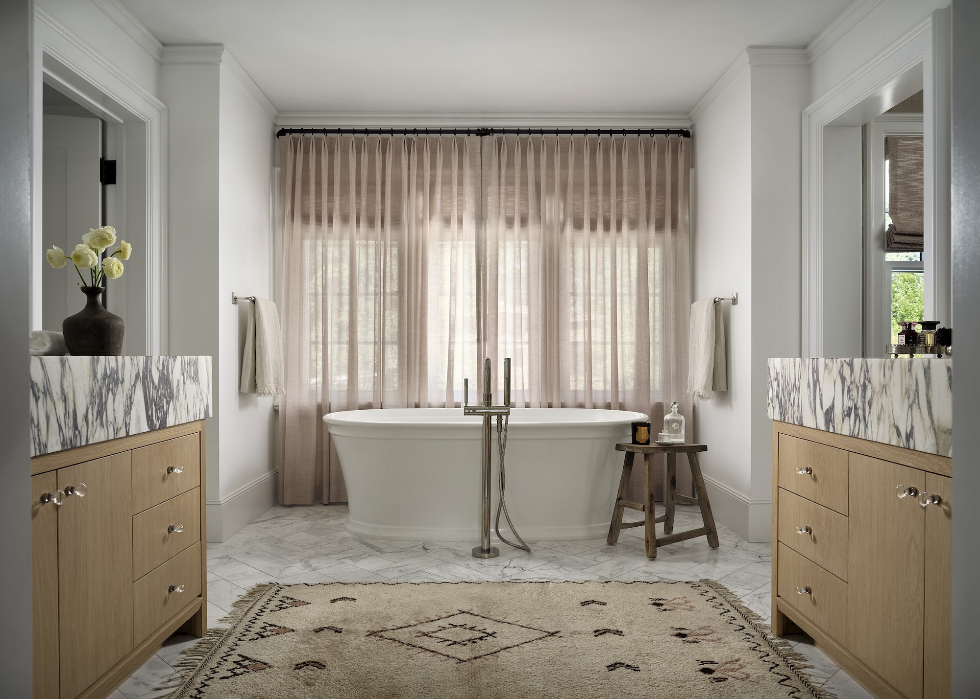 A large romantic style bathroom shown with a roll top bath, sheer linen curtains, a rug to soften the space and marble and wood vanity basins