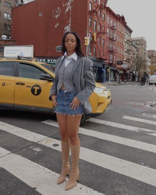 Woman wearing grey blazer, grey waistcoat, denim mini skirt and knee-high nude-colored boots while standing on New York street.