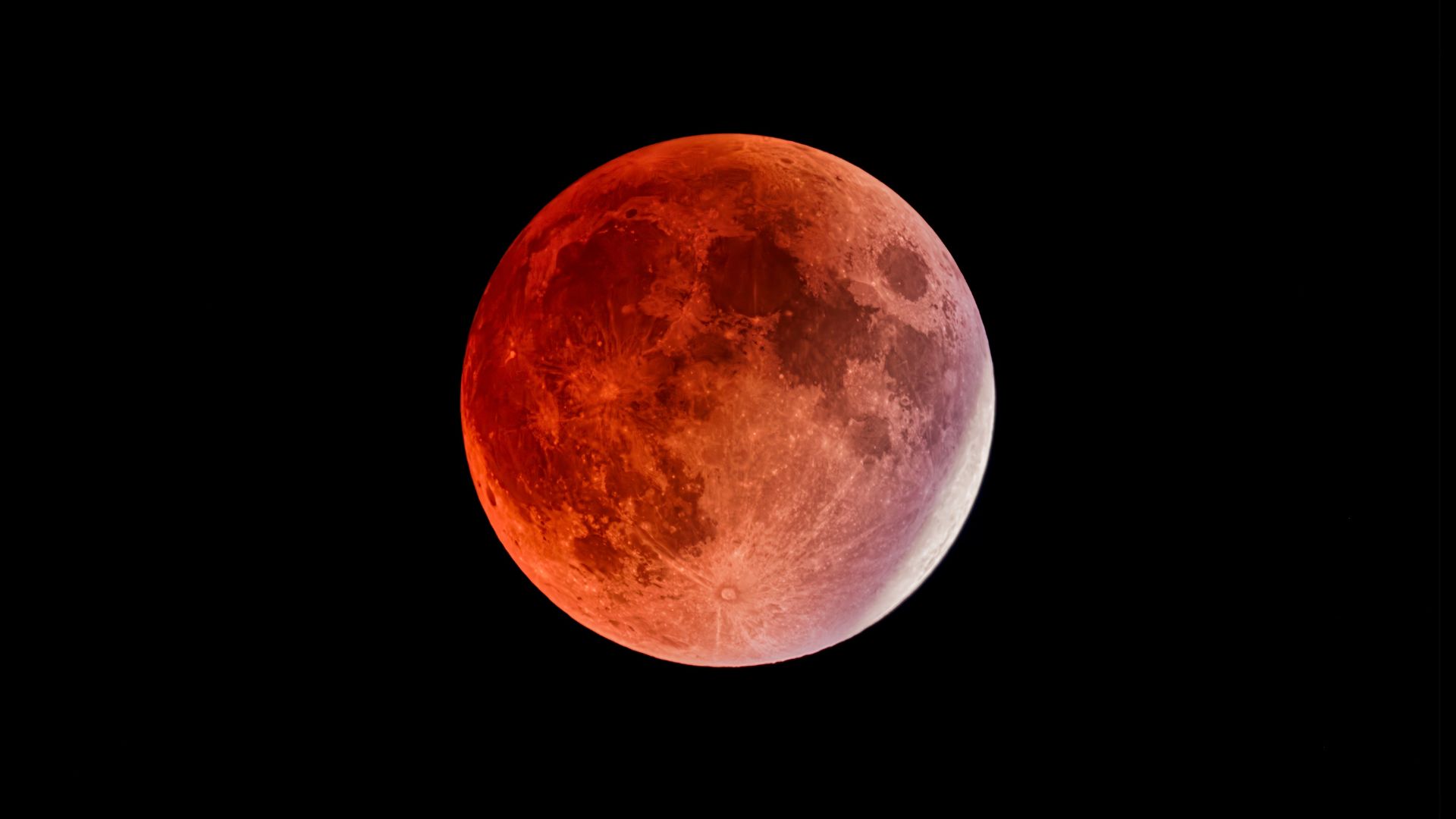 A full moon is photographed during a lunar eclipse in a black sky. The shadowed part of the lunarsurface is red and a thin crescent is exposed to direct sunlight on its right edge.