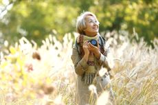 An older woman holding a camera and wearing a scarf looks up in awe in a field of wheat.