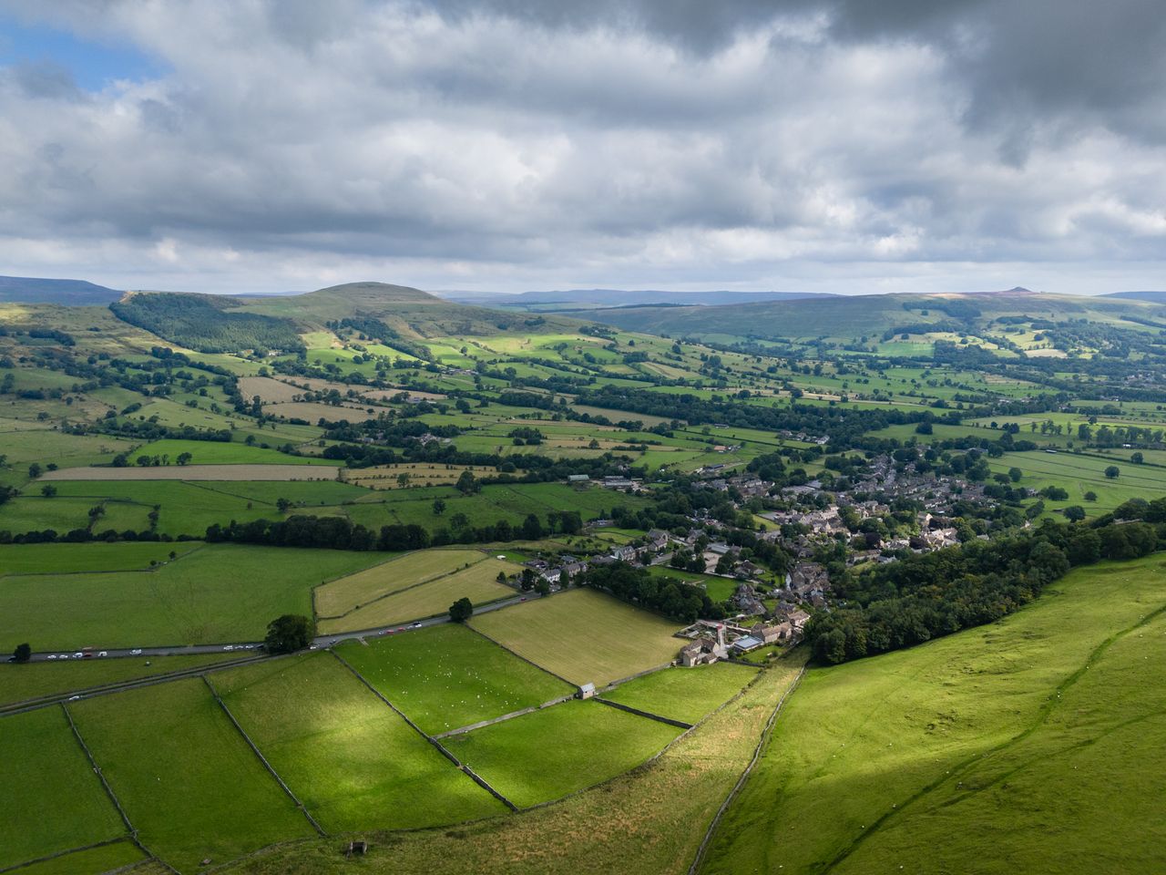 Image taken by DJI Air 3 (a camera drone). Here we see lots of green fields lined with hedgerows and a small town just to the right. In the distance are rolling green hills dotted with dark green trees and a gray, cloudy sky.