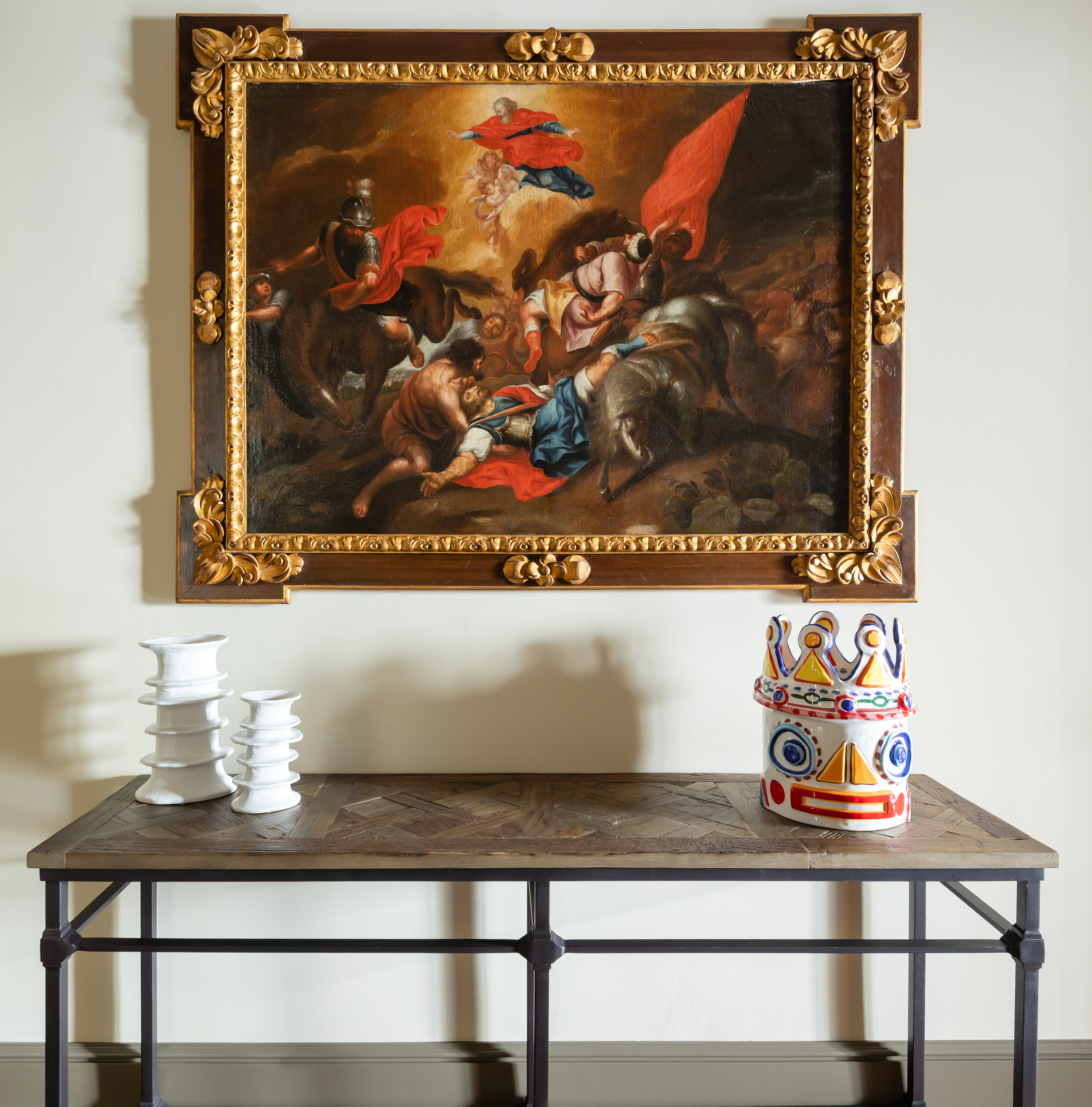 A figurative painting depicting a battle scene in bright tones is hung within a gold leaf-detailed wooden frame on a white wall on top of a wood and wrought iron table decorated with whimsical sculptures.