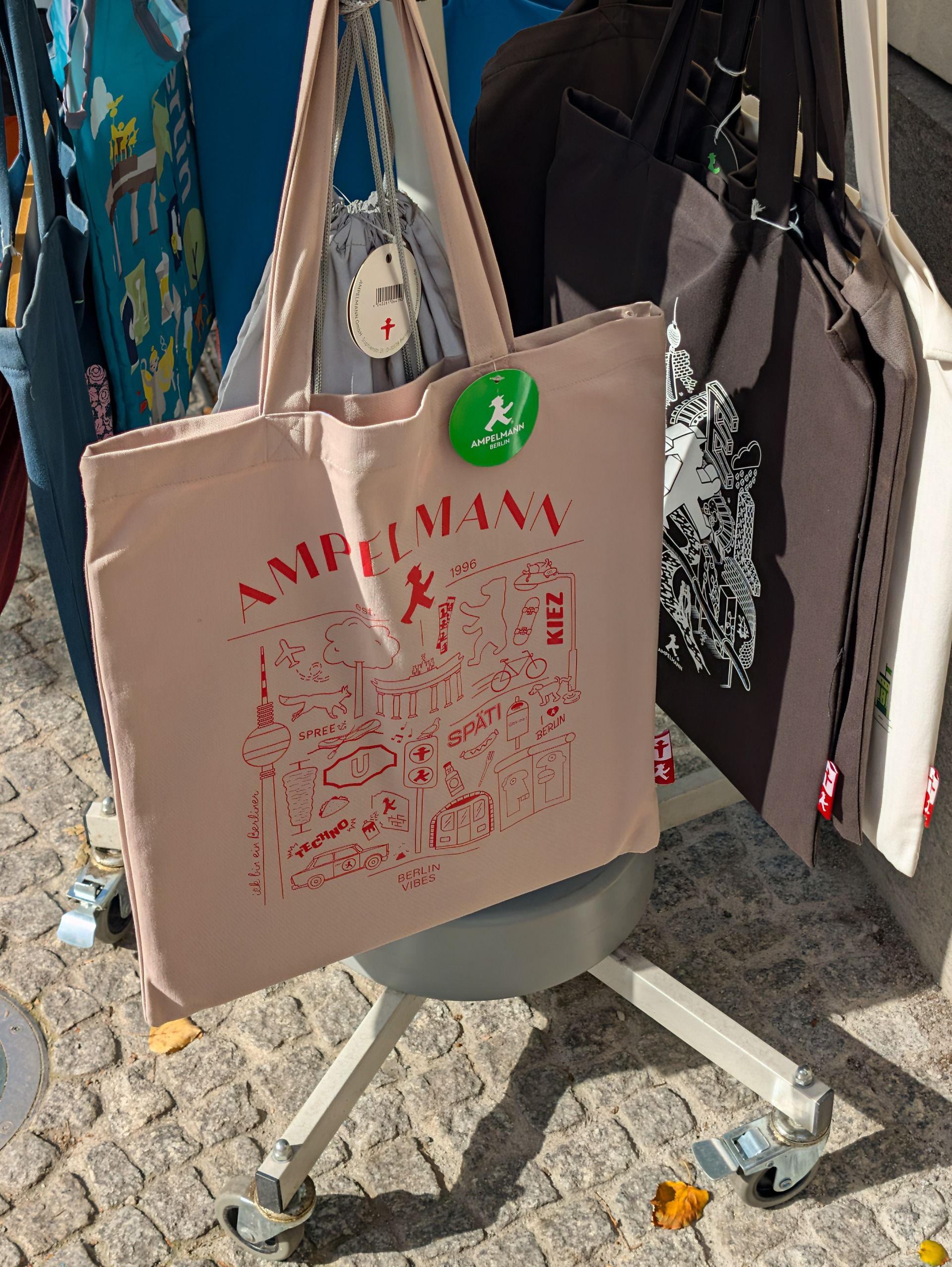 Beige and black Ampelmann Berlin tote bags hanging on an outdoor display stand.