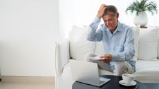 An older man looks surprised and confused as he looks at paperwork and his laptop while sitting on his living room sofa.