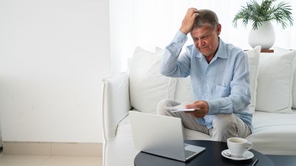 An older man looks surprised and confused as he looks at paperwork and his laptop while sitting on his living room sofa.