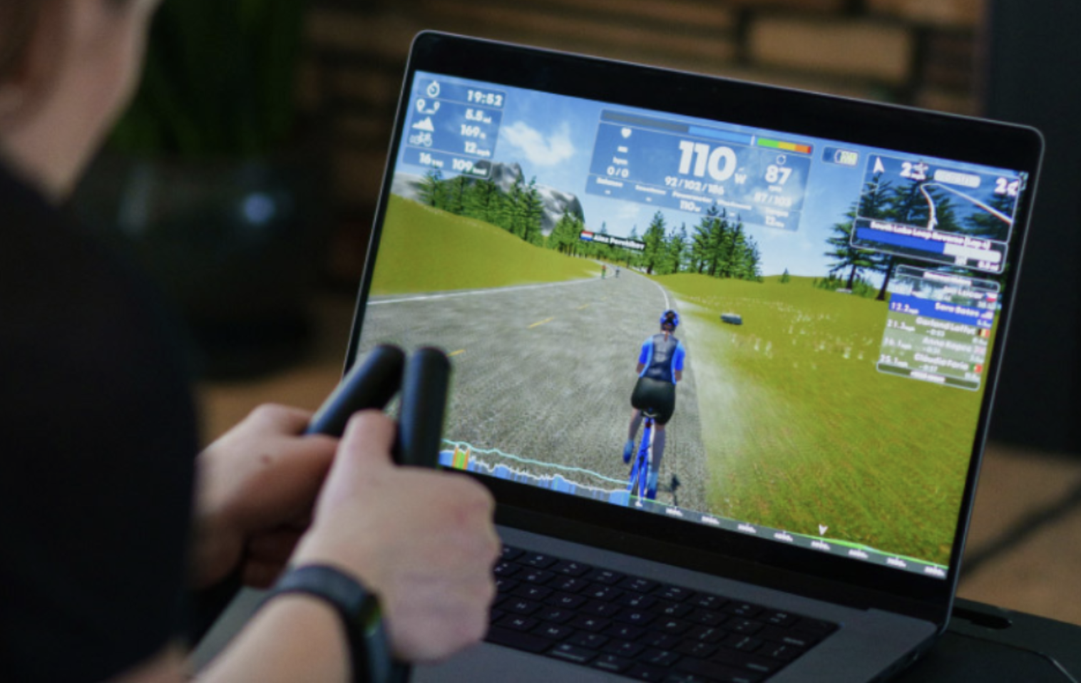 A view of a computer playing the racing simulation in front of a rider whose hands are only visible 