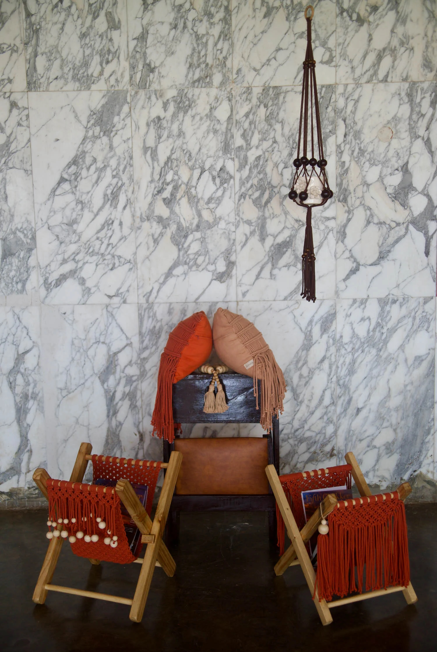 A series of furnishings, including two folding newspaper holders in red woven fibres and wood, and a leather and wood cabinet, sit in a room with dark floors and marble tiled walls.
