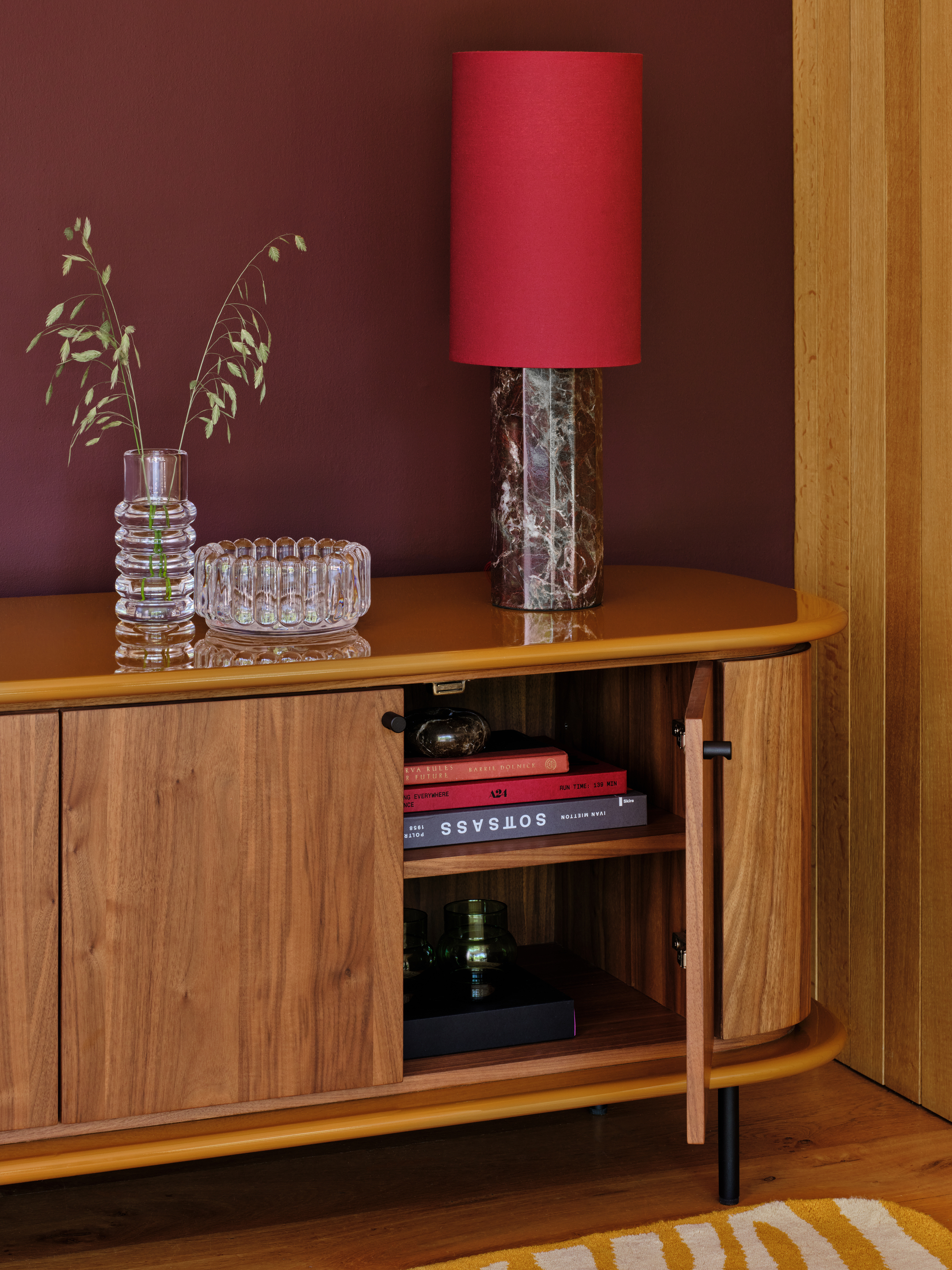 Image of a media console that is wooden with an ochre yellow lacquer top. There is a marble lamp with a red shade and two glass vases also on the console.
