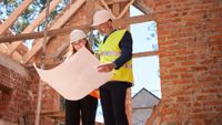 male and female wearing hi-vis and hard hats stood in partially built home looking at large set of plans