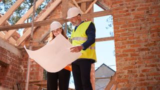 male and female wearing hi-vis and hard hats stood in partially built home looking at large set of plans