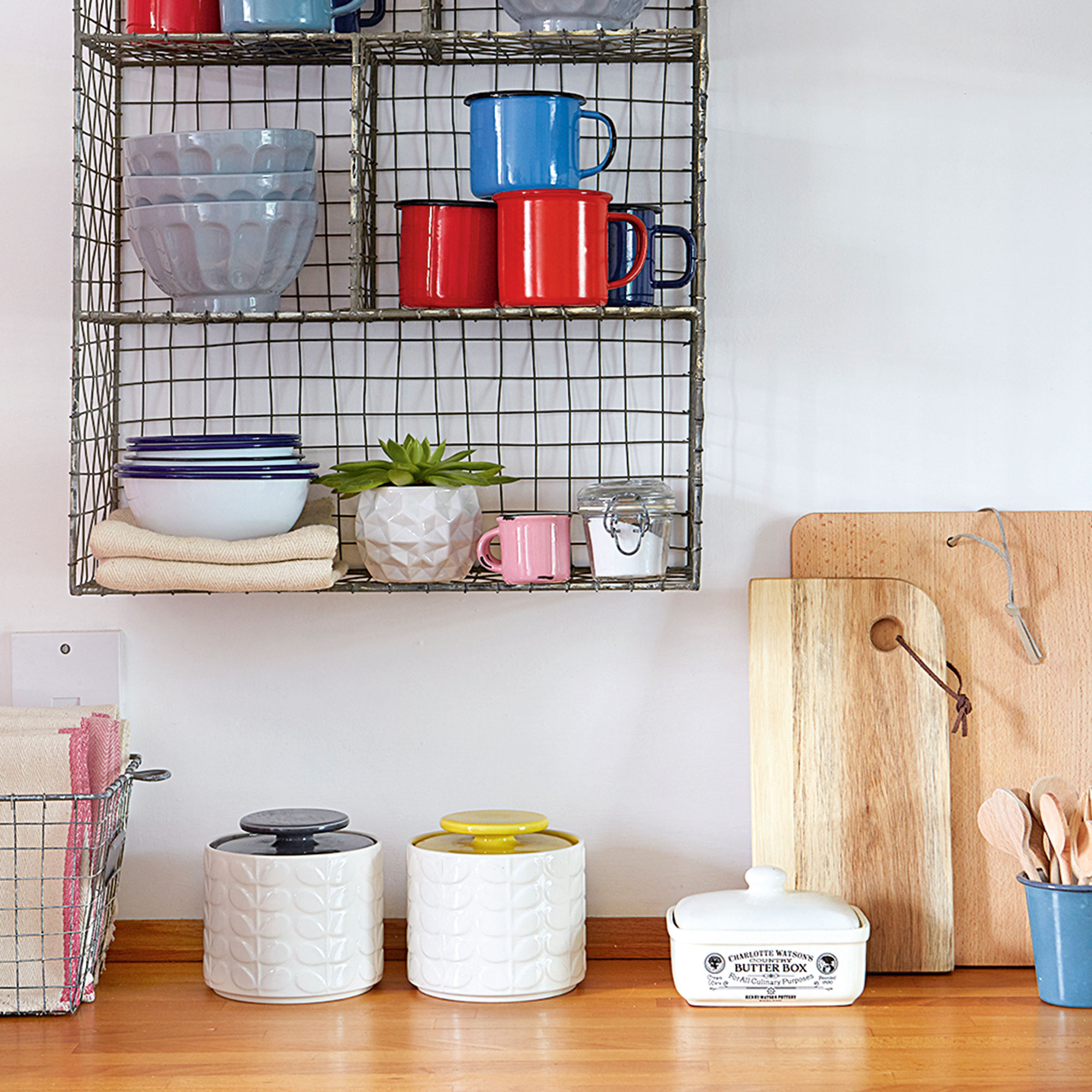 Ceramic storage jars and a ceramic butter dish sitting on a wooden kitchen worktop next to two propped up wooden chopping boards and below a wire wall shelf