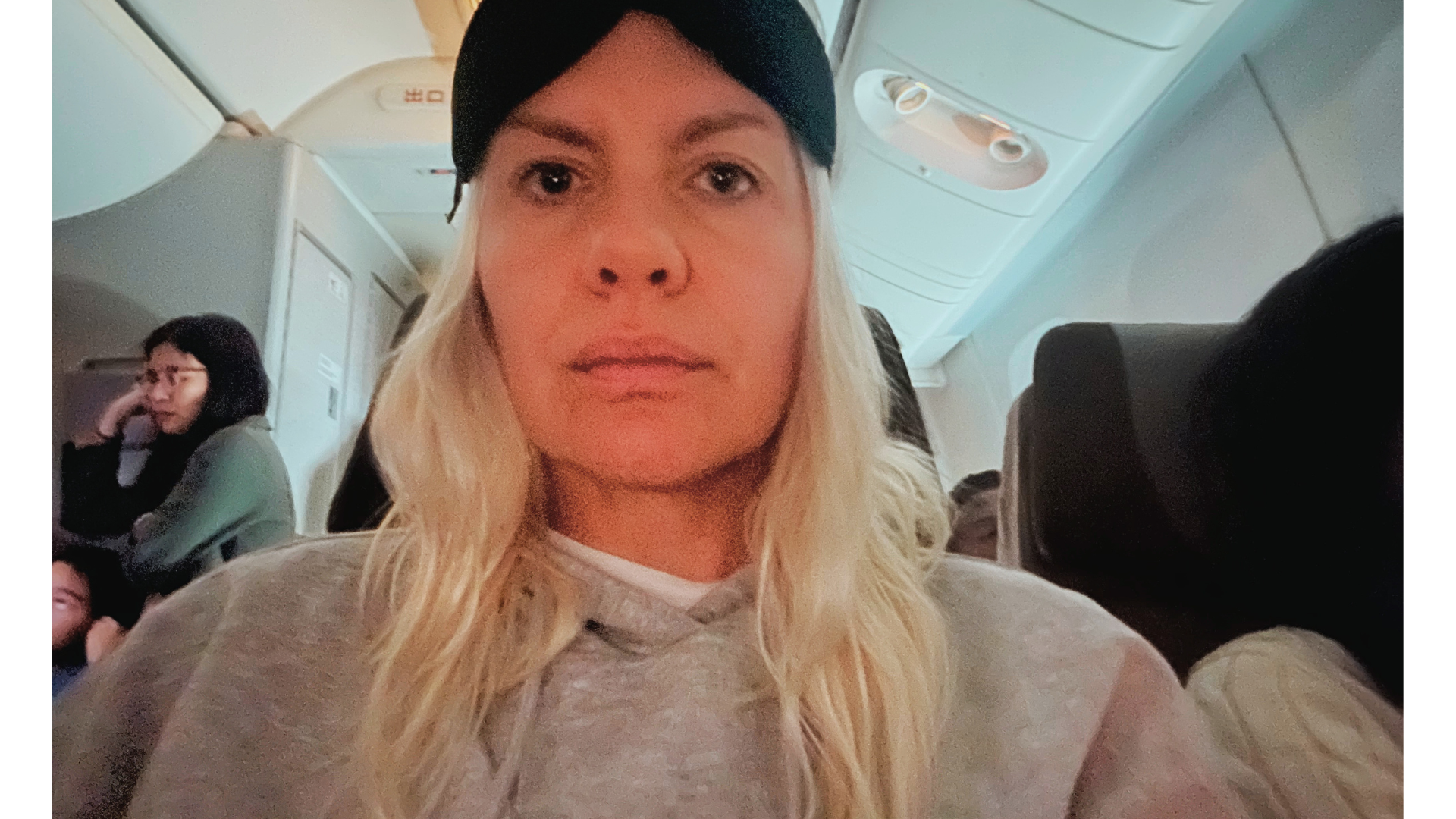 Jenny Haward wearing a grey hoodie on an airplane. She has an eye mask on the top of her head.