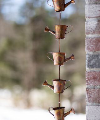 A copper rain chain with five watering can decorations, with a brick wall to the right of it and brown trees in the distance behind it