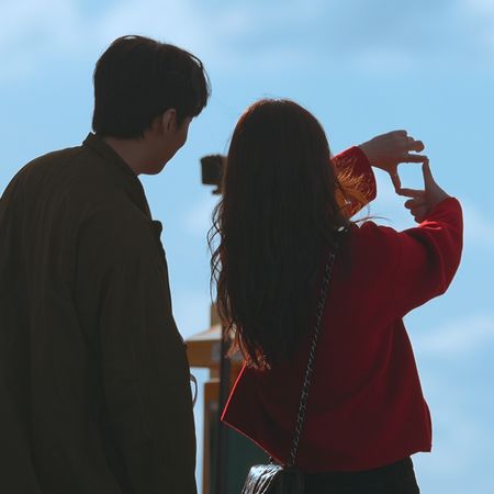 A man and woman are seen from the back as they stand under a blue sky. The woman is making a square with her fingers, in the still from Netflix's 'Better Late Than Single' season 2.