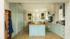 pale blue and off white kitchen with custom cabinetry, large kitchen island with wooden countertop and sink, wooden floorboards, ladder, artwork, view of kitchen table at far end, rug, white oven