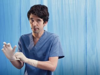 Ben Whishaw as Adam Kay in This Is Going To Hurt, standing in front of a blue hospital curtain wearing blue surgical scrubs and pulling on a pair of rubber surgical gloves