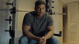 Alan Ritchson sits on a bench in front of a squat rack in a grey t-shirt and jeans in Reacher on Amazon Prime Video.