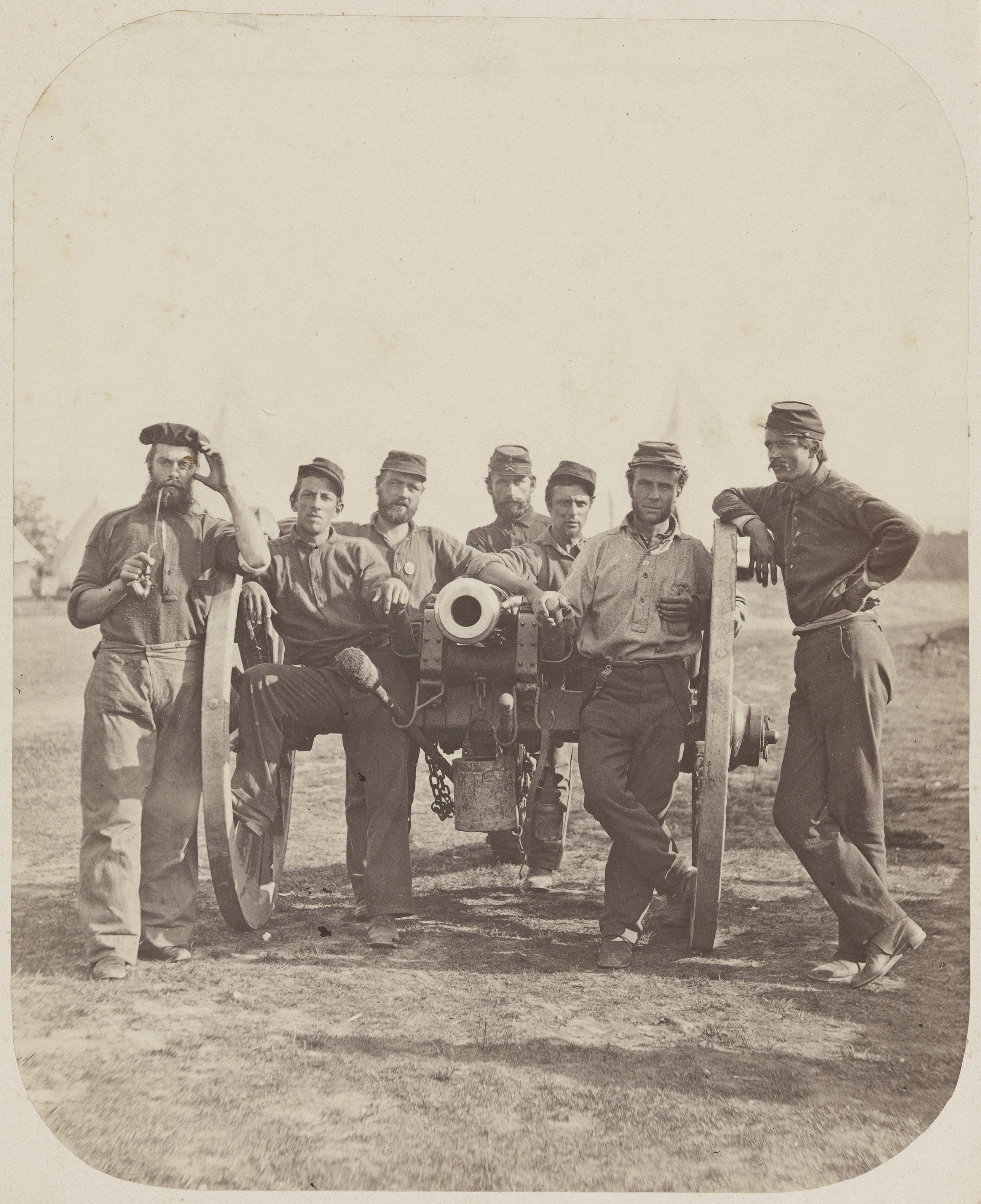 A group of seven Union soldiers in casual fatigue dress lean confidently against a large field artillery cannon in an open field.