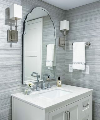 A bathroom with silver cubed wall sconces, an arched mirror, a white vanity with silver faucets, and a gray striped wallpaper