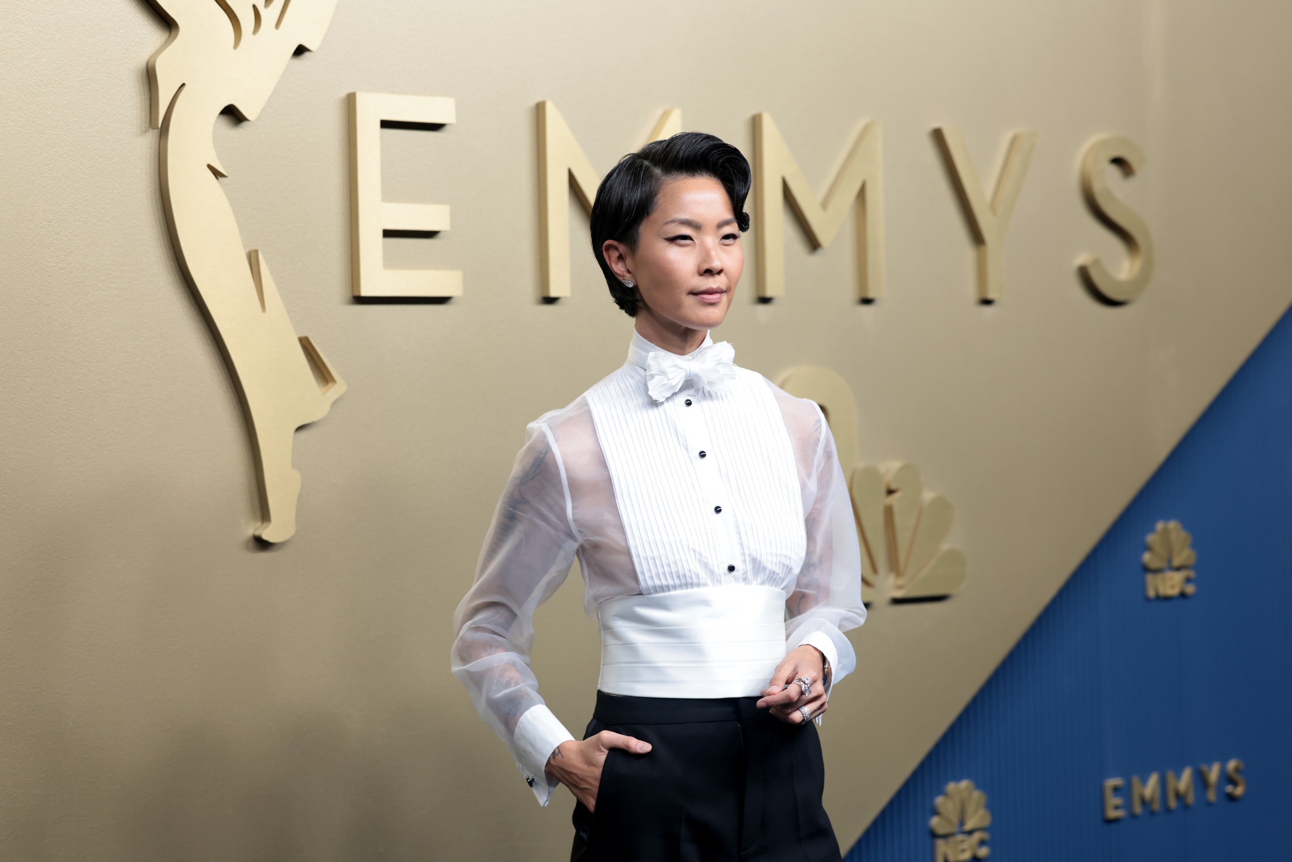 an asian woman in a tuxedo shirt and black pants in front of the gold emmys step and repeat