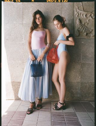 Models wearing a pink one-piece swimsuit and blue A-line skirt (left) and a blue one-piece swimsuit (right), both with black heeled sandals and mesh bags from the J.Crew x Maryam Nassir Zadeh collection.