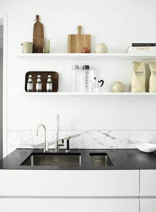 Image of a white kitchen with a black countertop and white shelving and cabinets. There are cutting boards, bottles, and bags of food on the shelving.