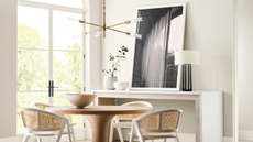 A beige dining room with high ceilings, glass doors, a round wooden table and a large framed photograph resting on a side table
