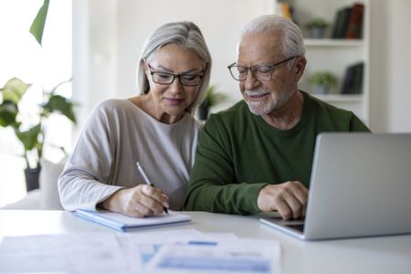 Senior couple sitting at home, using a laptop and taking notes while reviewing household finances, budgeting and online information together.