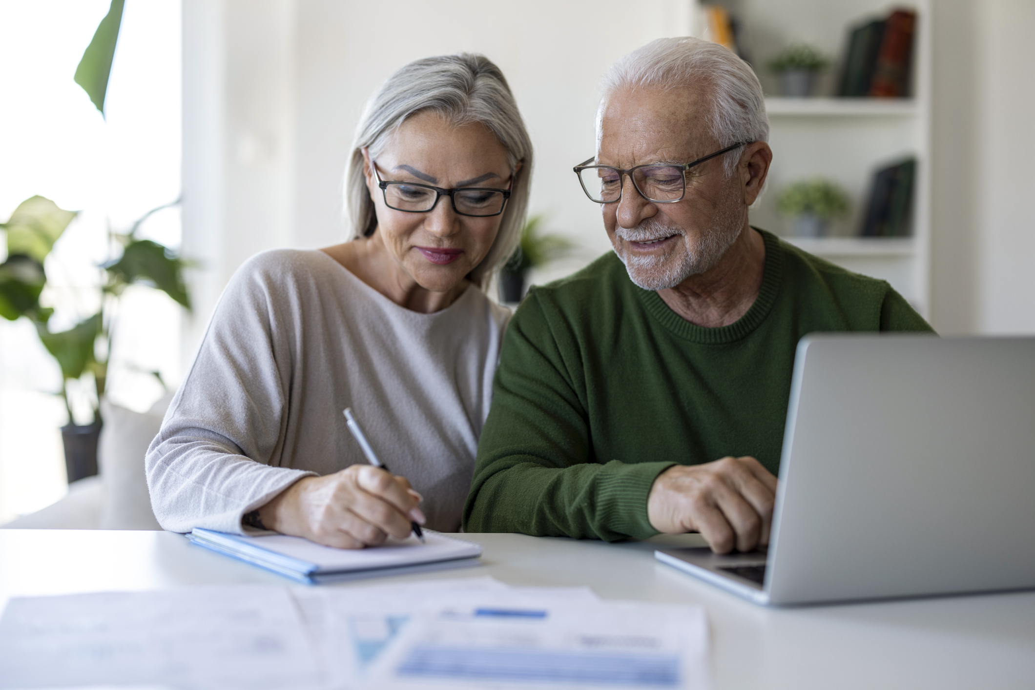 Senior couple sitting at home, using a laptop and taking notes while reviewing household finances, budgeting and online information together.