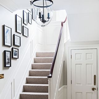 A white hallway with a picture gallery going up the staircase next to white door and pendant light