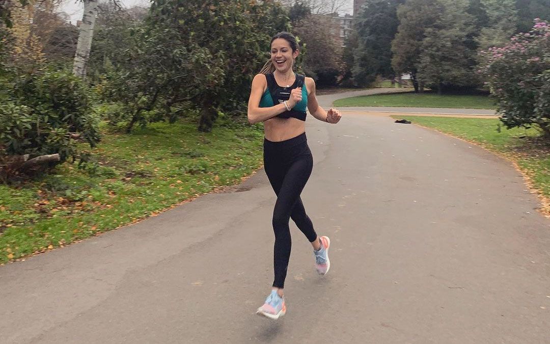 A woman runs on a Tarmac path in a park. She is smiling and wearing black leggings and a sports bra.