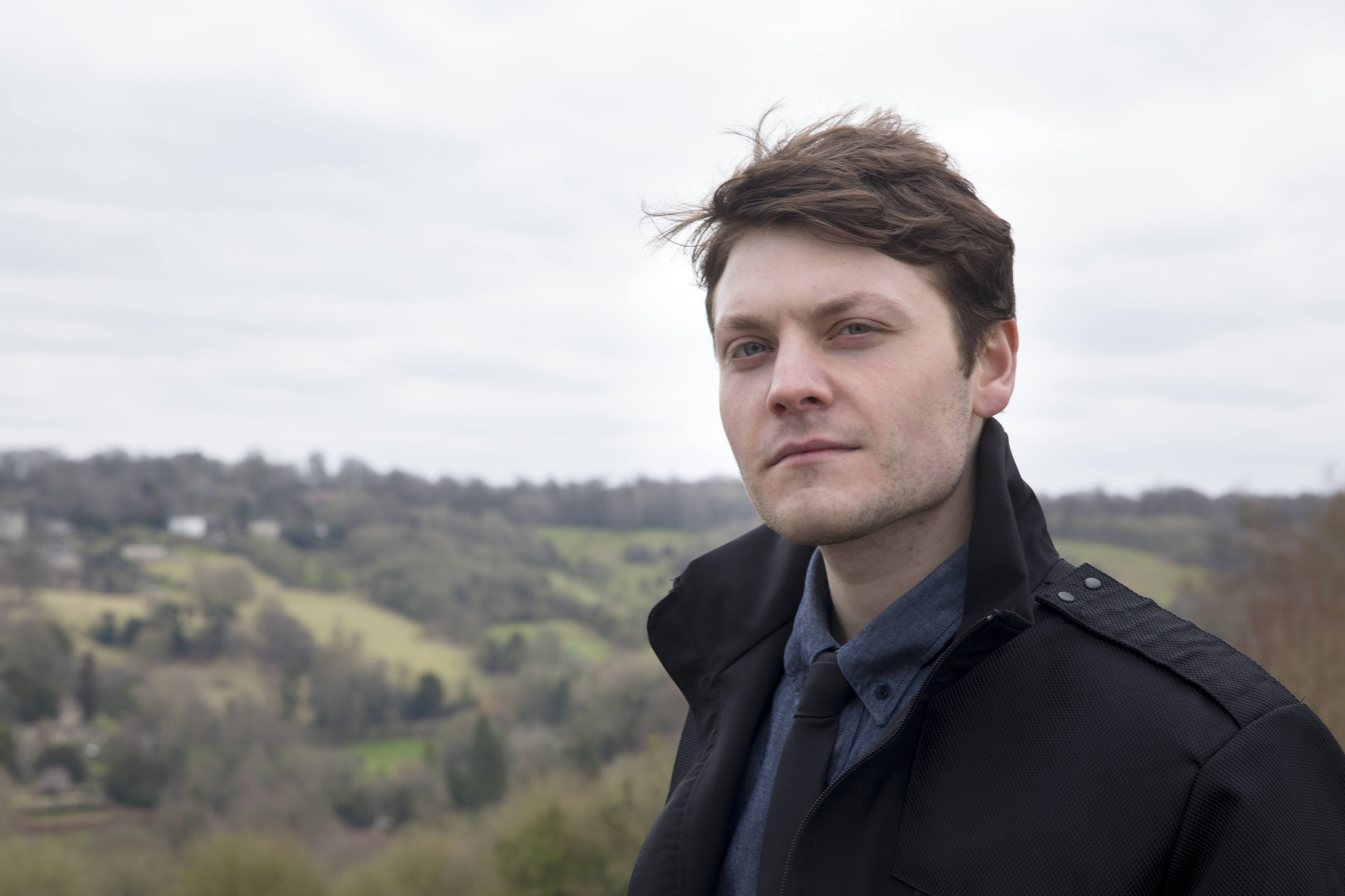 White man with dark hair, shirt and tie and black jacket, posing against a backdrop of a hillside and sky