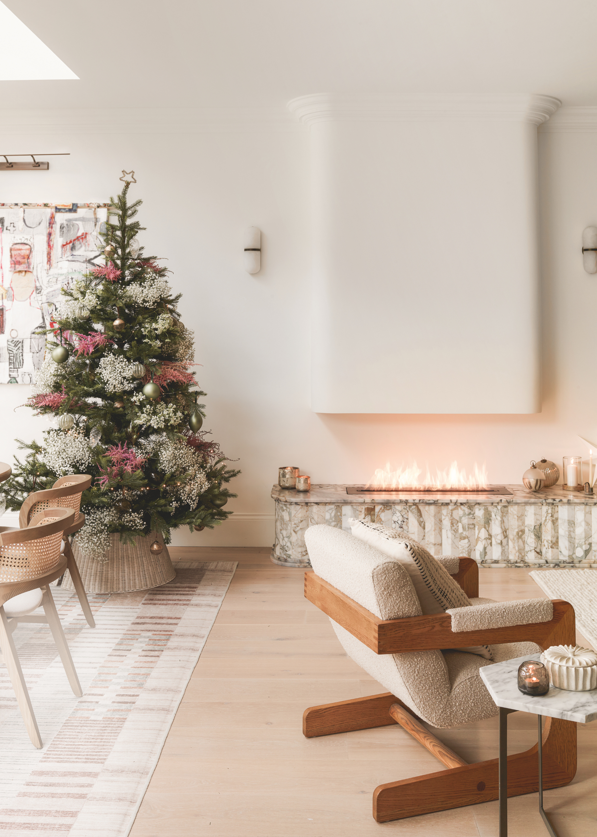 a modern, neutral living space decorated for christmas with a christmas tree in the corner a modern tabletop fireplace lit against the back wall and a boucle armchair with a wooden frame