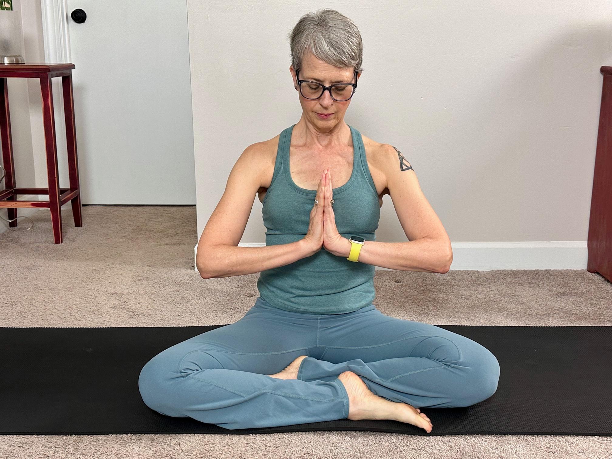 woman wearing blue leggings and vest sat cross legged with hands in prayer position on a black exercise mat on carpet indoors