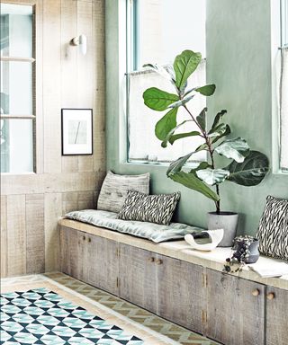 Green hallway with textured, green painted walls, wooden wall paneling and custom, built in wooden bench, accessorized with plants cushions, framed wall art, geometric blue and black rug