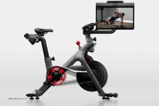 The Peloton Cross Training Bike + in full from the side with the screen showing a white man completing a devel press with dumb bells 