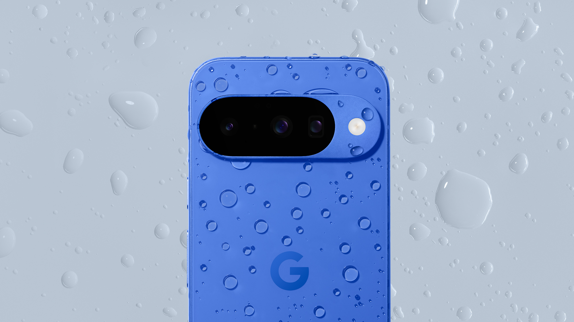 An indigo blue version of the Google Pixel 10 phone covered in water droplets on a grey background