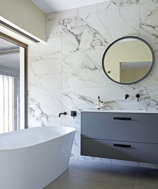 bathroom with freestanding tub, marble finish wall tiles, gray vanity, round mirror