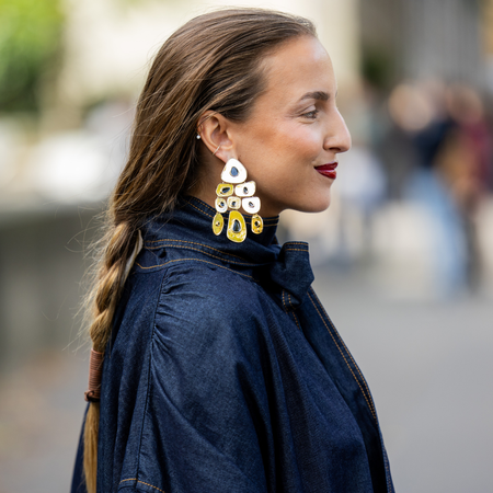 A side on view of a guest at paris fashion week wearing statement earrings, a red lip and a low plait