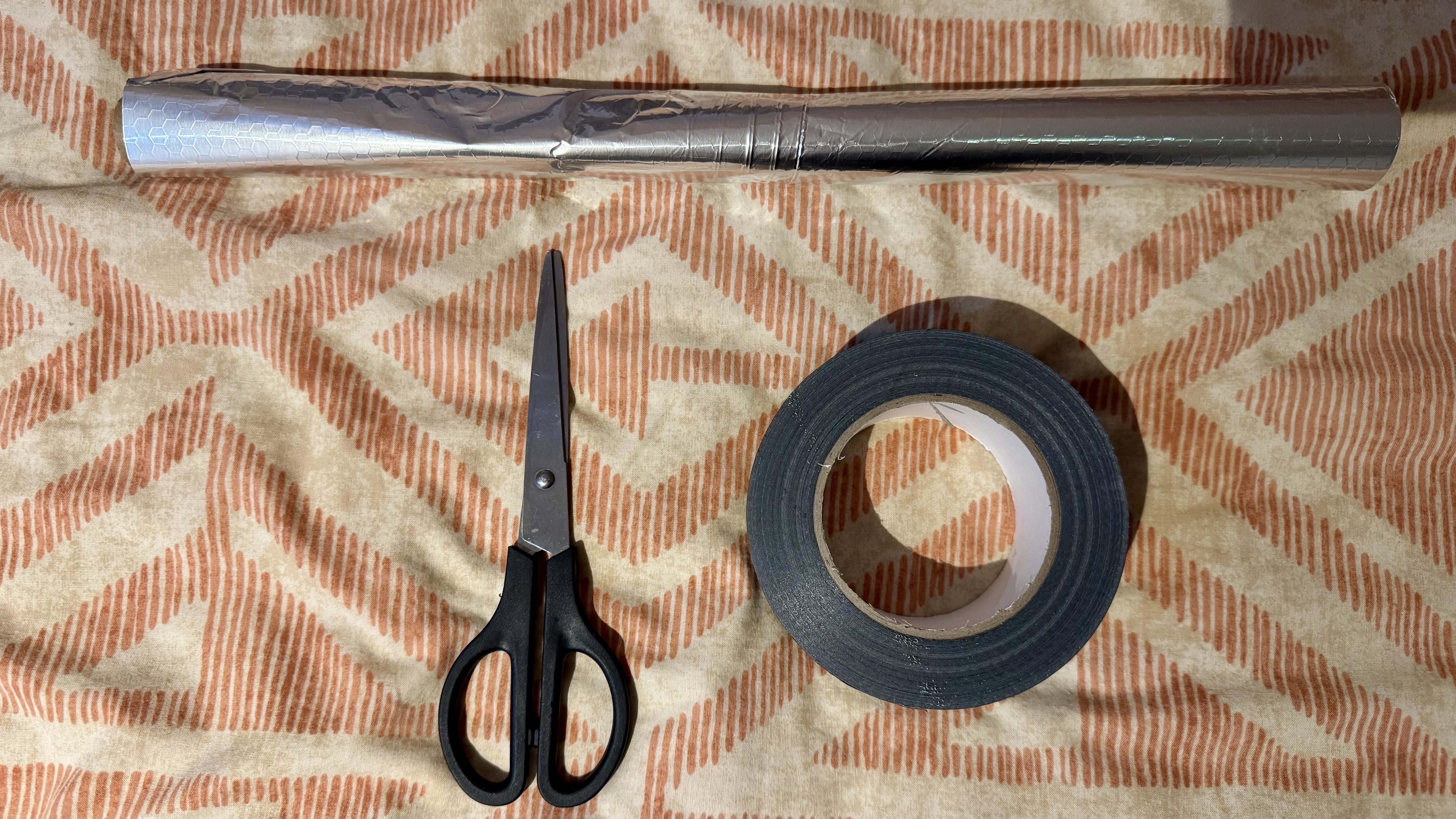 A roll of tin foil, black duct tape and scissors on orange patterned bedding
